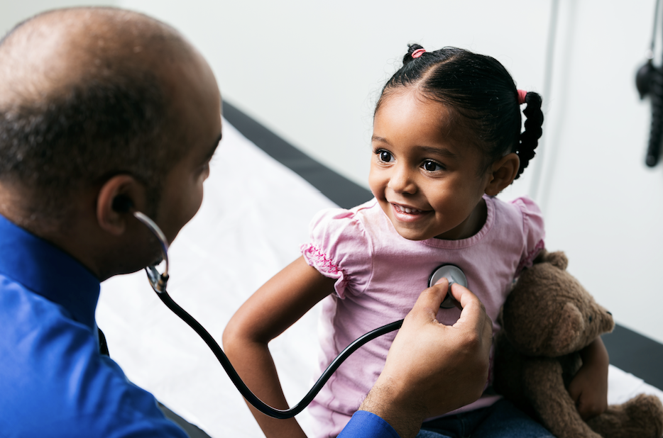 A healthcare professional uses a stethoscope to listen to a smiling child's heartbeat while she holds a teddy bear.